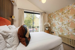A hotel room with a bed, a window with curtains, a radiator, and a wall with a floral pattern. - Riverside Fulham Living | Stunning Elegant London Home Steps from the Thames (London)
