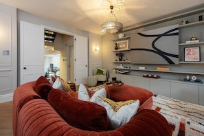 A living room with a red sofa, a chandelier, and a wall with shelves. - Riverside Fulham Living | Stunning Elegant London Home Steps from the Thames (London)
