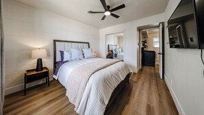 Une chambre à coucher comprenant un lit, une table de chevet, une lampe, un ventilateur de plafond et un téléviseur fixé au mur.