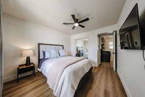 Ein Schlafzimmer mit Bett, Nachttisch, Lampe, Deckenventilator und einem an der Wand montierten Fernseher.