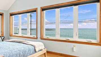 A bedroom with a bed, two windows with wooden frames, and a view of the sea.