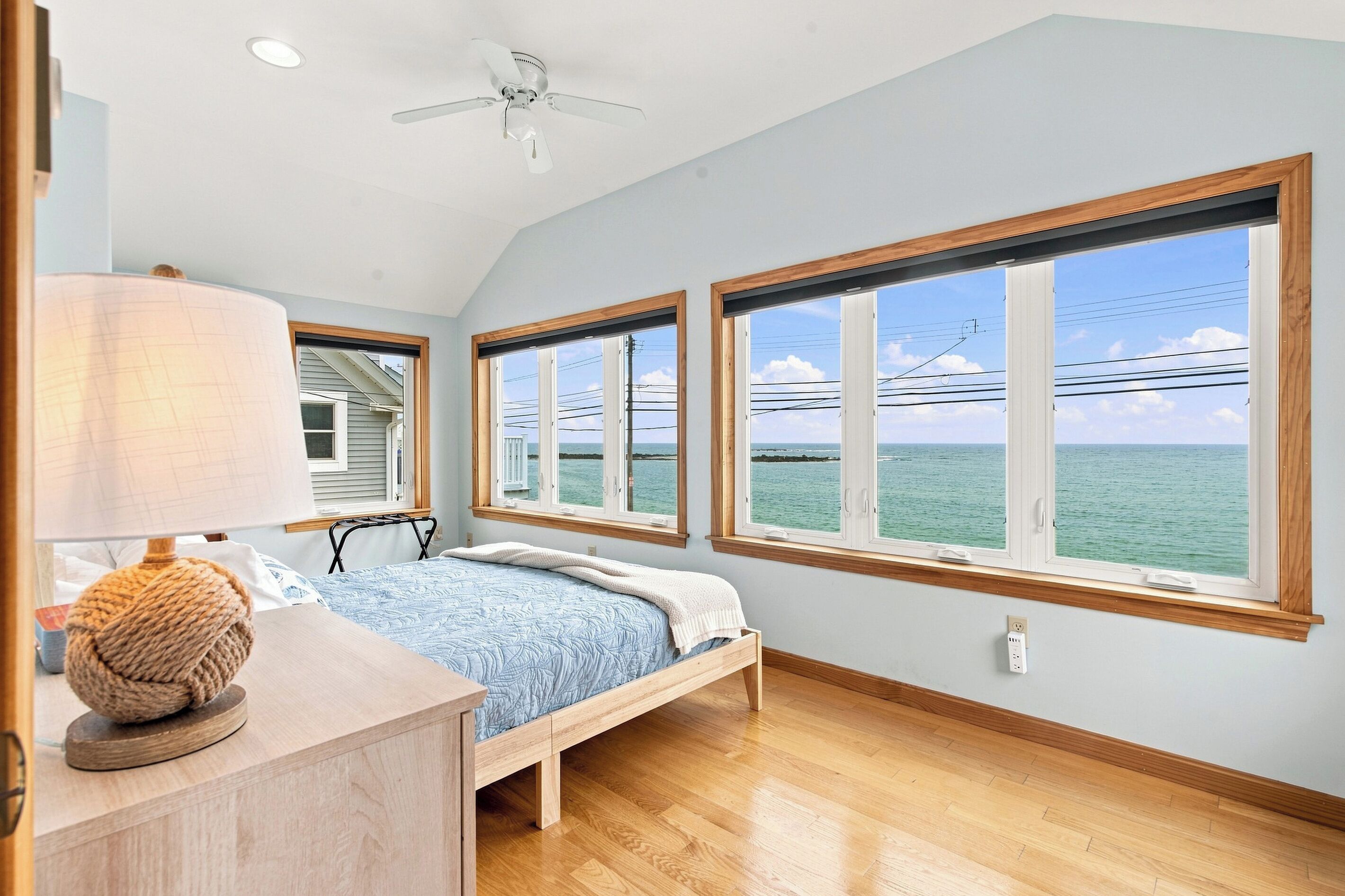 A bedroom with a bed, a bedside table with a lamp, and large windows with a view of the sea.