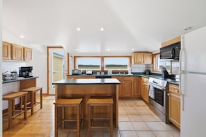 A modern kitchen with wooden cabinets, a large island with bar stools, a built-in oven, and a view of the outdoors. - Wells Home with Ocean & Marsh Views + Beach Access (Wells)
