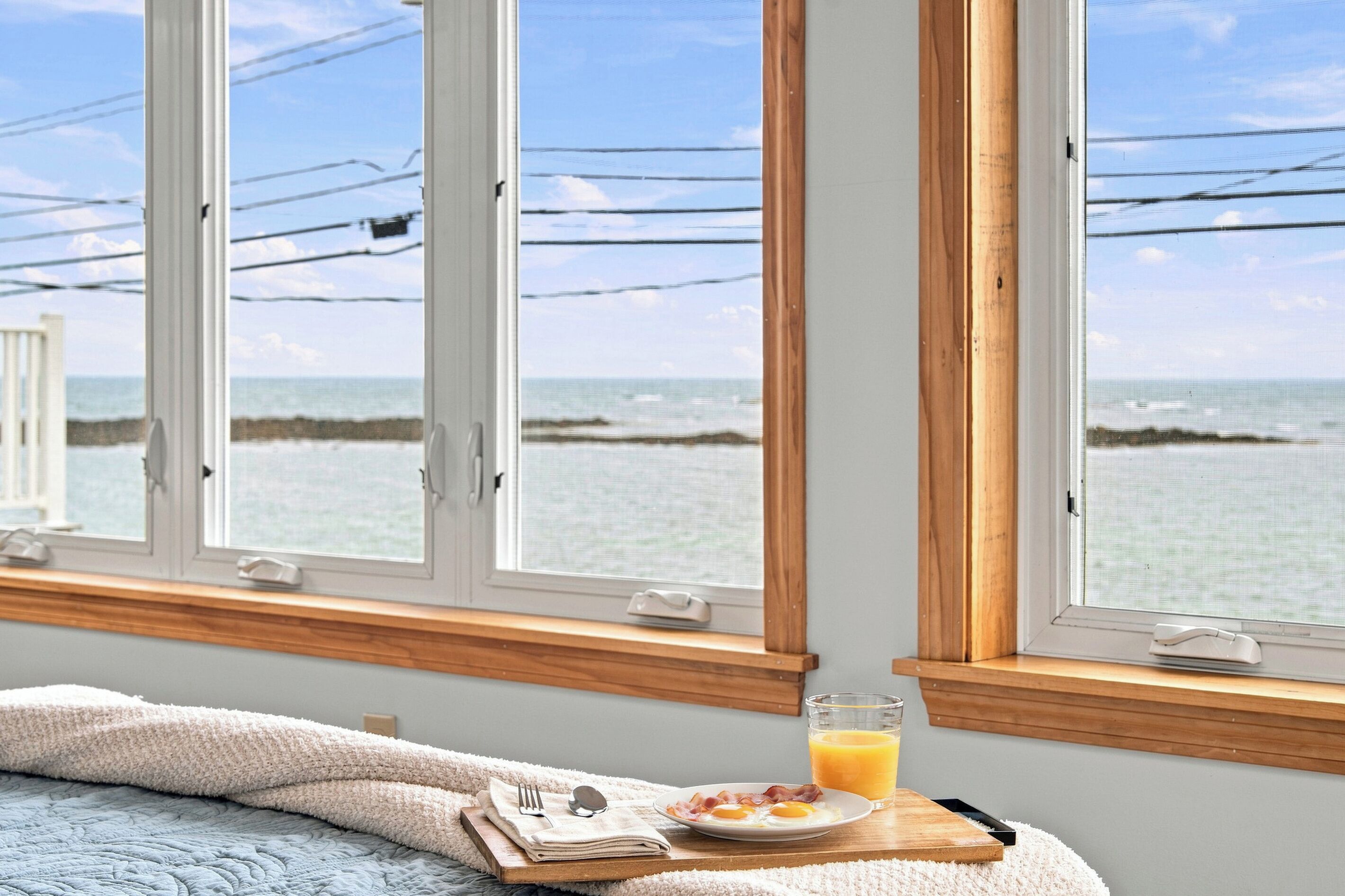 Un dormitorio con vistas al océano a través de ventanas amplias, una cama con manta blanca, una bandeja de desayuno con huevos y jugo de naranja, y una mesita de noche con un vaso.