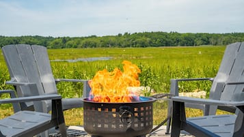 Two Adirondack chairs facing a campfire with a grassy field and trees in the background.