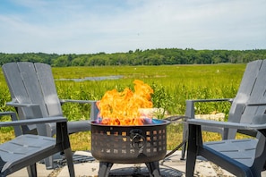Two Adirondack chairs facing a campfire with a grassy field and trees in the background. - Wells Home with Ocean & Marsh Views + Beach Access (Wells)