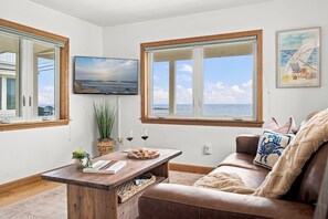 A living room with a wooden table, a sofa, a television, and a view of the sea. - Wells Home with Ocean & Marsh Views + Beach Access (Wells)