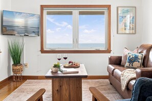 A living room with a large window, a flat-screen TV, a wooden table with a book and glasses, a brown leather sofa, and a potted plant. - Wells Home with Ocean & Marsh Views + Beach Access (Wells)