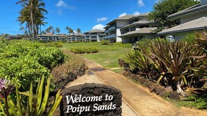 Een pad dat leidt naar een hotel met een bord waarop staat 'Welcome to Poipu Sands', omringd door tropische planten en palmbomen.