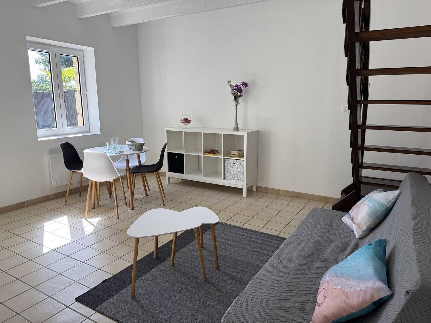 Uma sala de jantar com mesa e cadeiras, uma mesa consola branca com objetos decorativos, um sofá cinzento com almofadas, e uma escada.