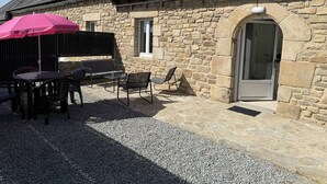 Un edificio de piedra con techo inclinado, un patio con mesas al aire libre y una puerta con panel de vidrio.