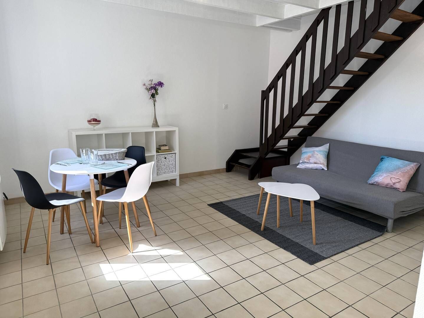Un salon moderne comprenant une table à manger, des chaises, un canapé, un escalier et une console blanche ornée d’un vase.