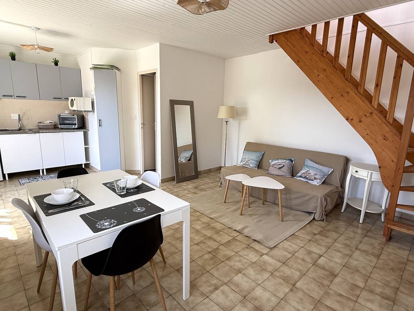 A modern kitchen with white cabinets, a dining table with chairs, a sofa with cushions, a mirror, and a wooden staircase.