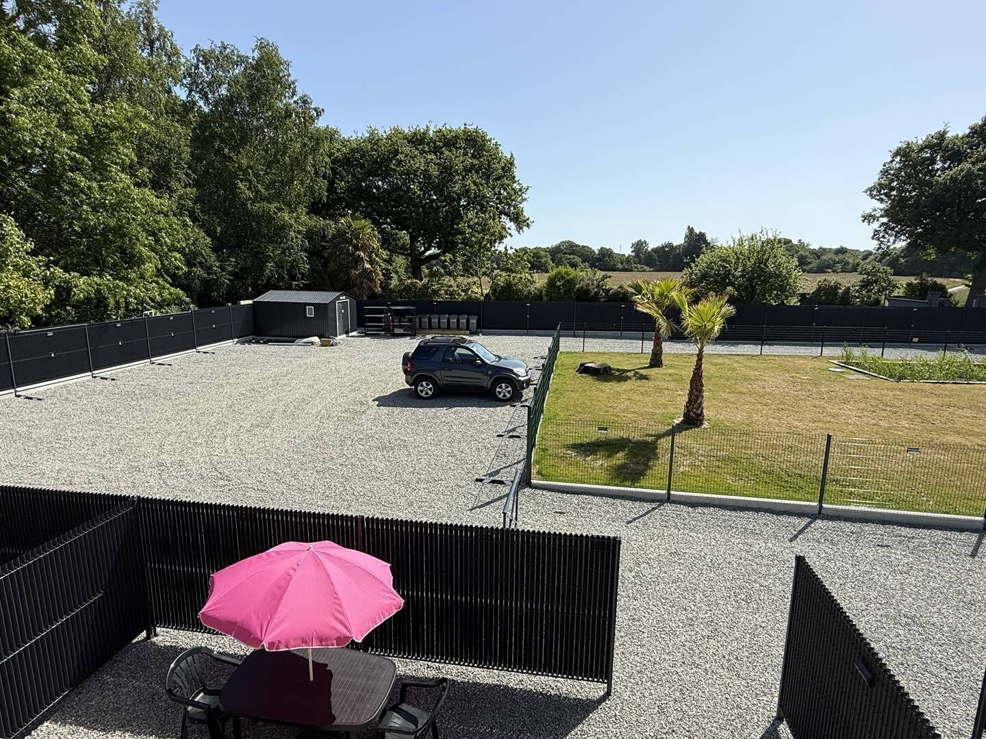 A gravel parking area with a car, a small shed, and a fenced grassy area with palm trees.