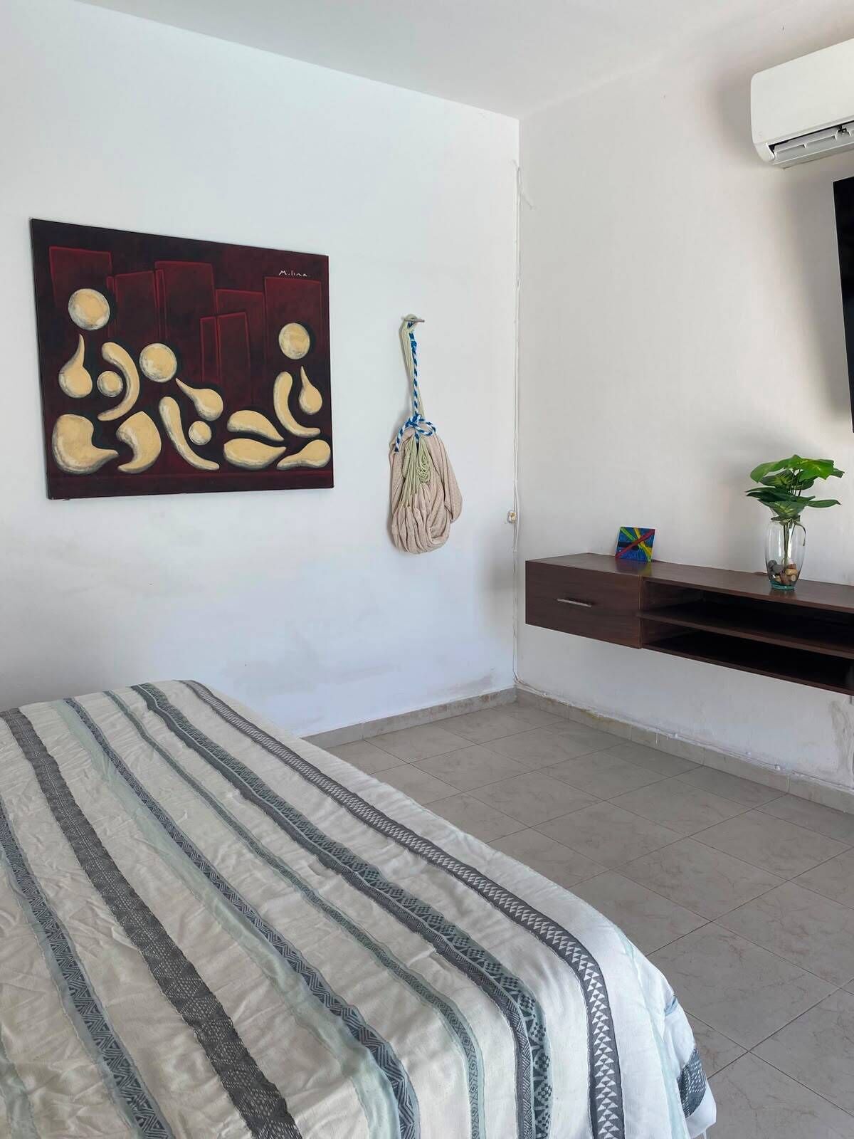 A bedroom with a bed, a wall-mounted air conditioner, a painting, a hanging bag, a small plant, and a shelf with a vase.