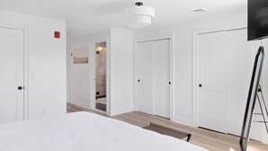 Un dormitorio con cama, paredes blancas, un televisor de pantalla plana y una puerta que da a un baño.