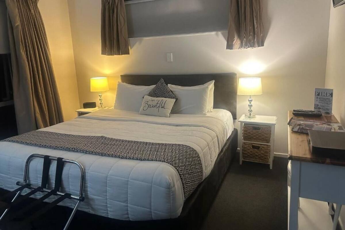 Habitación de hotel con cama, dos lámparas de noche, mesita de noche, escritorio con silla y cortinas.