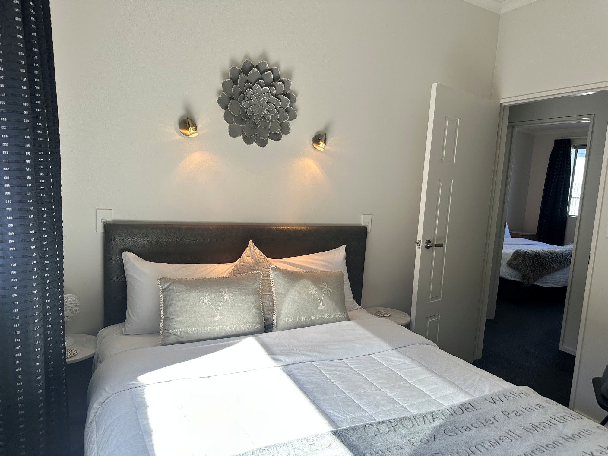 Una cama bien tendida con una colcha blanca y almohadas, un aplique de pared y una puerta que da a otra habitación.