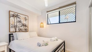 A bedroom with a bed, a bedside table, a wall-mounted picture, a window with a view, and a small wall light. - Sea Air Number Two On Queen (Yeppoon)