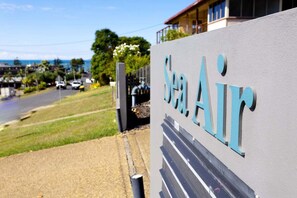 Ein Schild mit der Aufschrift 'Sea Air' an einem Gebäude, eine Straße mit Fahrzeugen und im Hintergrund Meer und Grün.