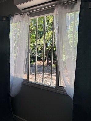 Ein Fenster mit transparenten weißen Vorhängen, Blick auf Bäume und Kiesgrund draußen, sowie ein Deckenlüfter.