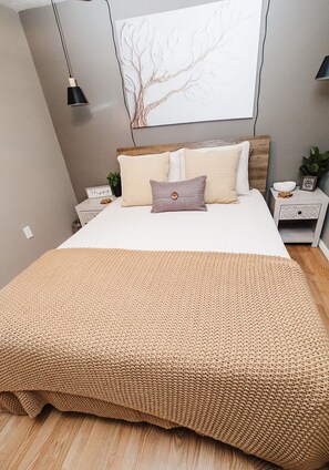 Ein Schlafzimmer mit Bett, Kissen, Nachttisch und einer Wandlampe, außerdem eine Pflanze.