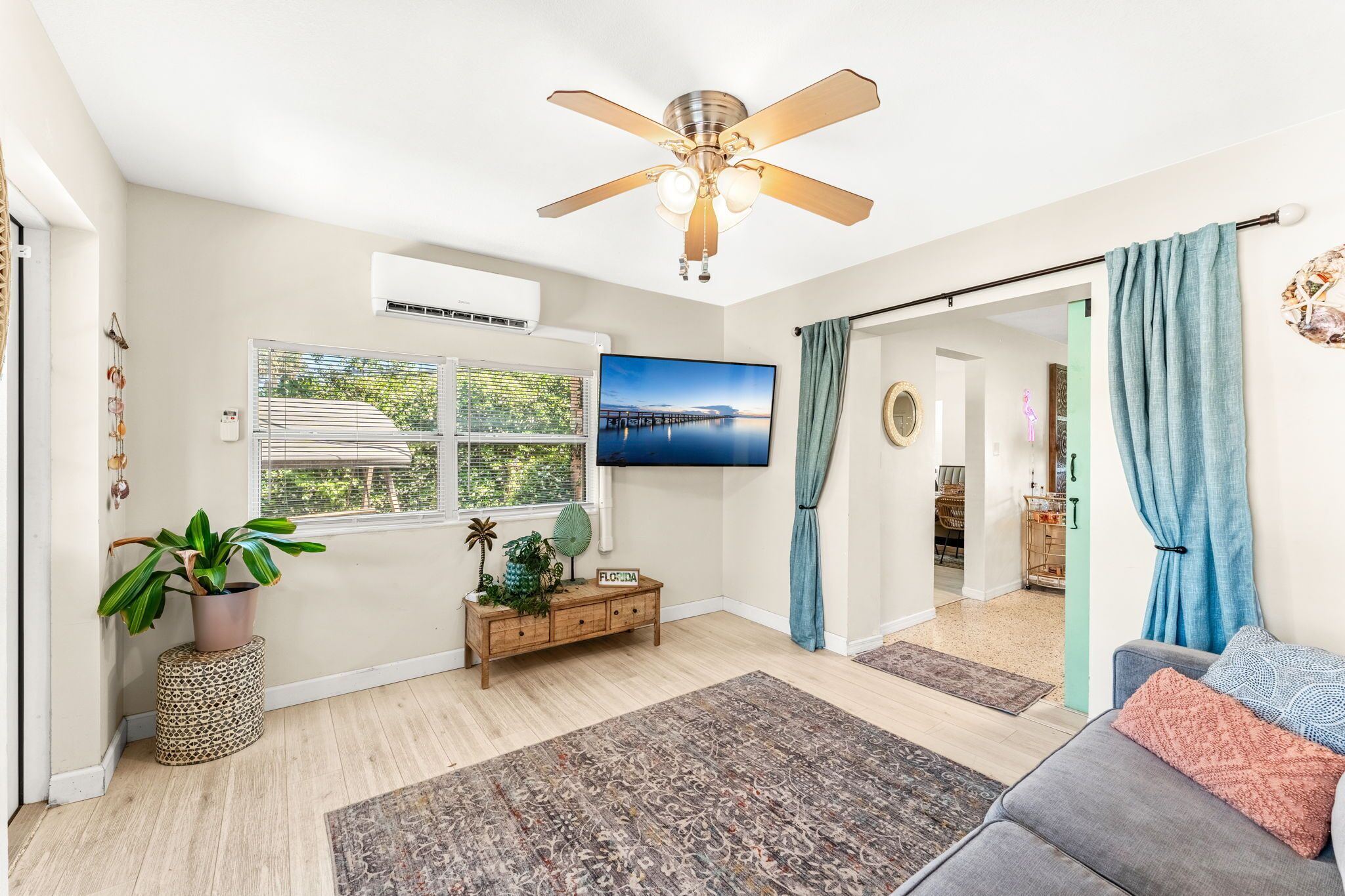 A living room with a ceiling fan, a flat-screen TV, a window with blinds, a potted plant, a wooden console table, and a gray sofa.