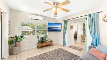 A living room with a ceiling fan, a flat-screen TV, a window with blinds, a potted plant, a wooden console table, and a gray sofa.