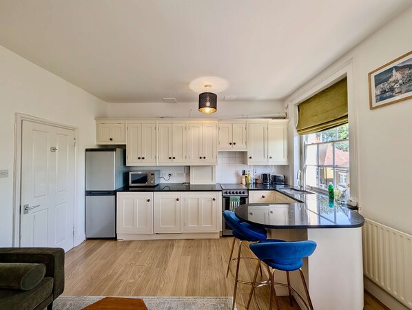 A kitchen with white cabinets, a black countertop, a microwave, a stove, and a sink. There are two blue bar stools and a window with a view of a building. - Two bed Flat Near King s Cross St Pancras (London)