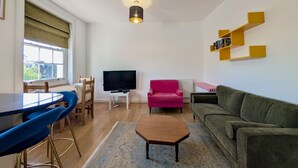 Una sala de estar con televisor, un sillón rosa, un sofá verde, una mesa de centro de madera y una estantería amarilla.