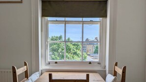 Una habitación con una mesa y sillas de madera, una ventana con vistas a árboles y edificios, y un radiador en la pared.
