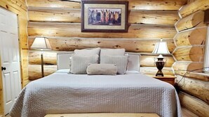 Ein Schlafzimmer in einer Blockhütte mit einem Bett, Kissen, einer Nachttischlampe und einem gerahmten Bild an der Wand.