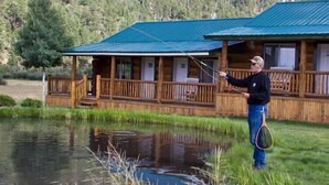 Eine Holzhütte mit grüner Dachdeckung, ein Steg, der über einen Teich führt, und eine Person beim Angeln.