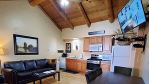 A living room with a black leather sofa, a dining table, and a kitchen area with wooden cabinets and a white refrigerator. - Knotted Pine Suite #251 (Greer)