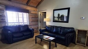 A log cabin interior with two leather sofas, a coffee table, a framed picture on the wall, and a window with blinds. - Knotted Pine Suite #251 (Greer)