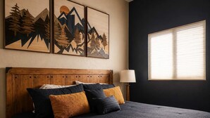 Un dormitorio con cabecera de madera, cama con ropa de cama oscura, varios almohadones, una lámpara de noche y una ventana con persianas.