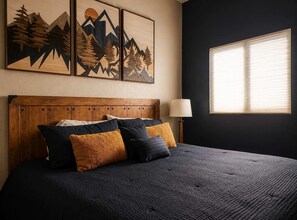 A bedroom with a wooden headboard, a bed with dark bedding, multiple pillows, a bedside lamp, and a window with blinds.