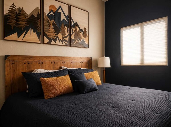 A bedroom with a wooden headboard, a bed with dark bedding, multiple pillows, a bedside lamp, and a window with blinds.