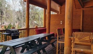 Een houten terras met een picknicktafel en stoelen, een barbecue en uitzicht op bomen.