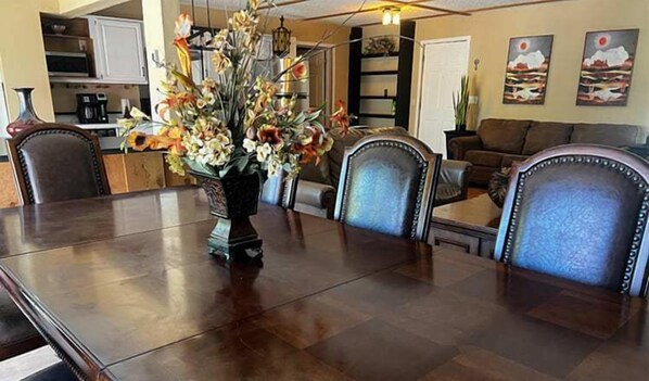 A dining area with a large wooden table, chairs, a vase of flowers, and a kitchen in the background.