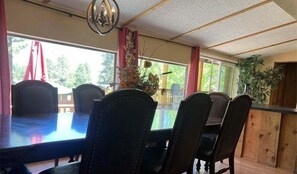 A dining area with a long table, chairs, a chandelier, and large windows with curtains.