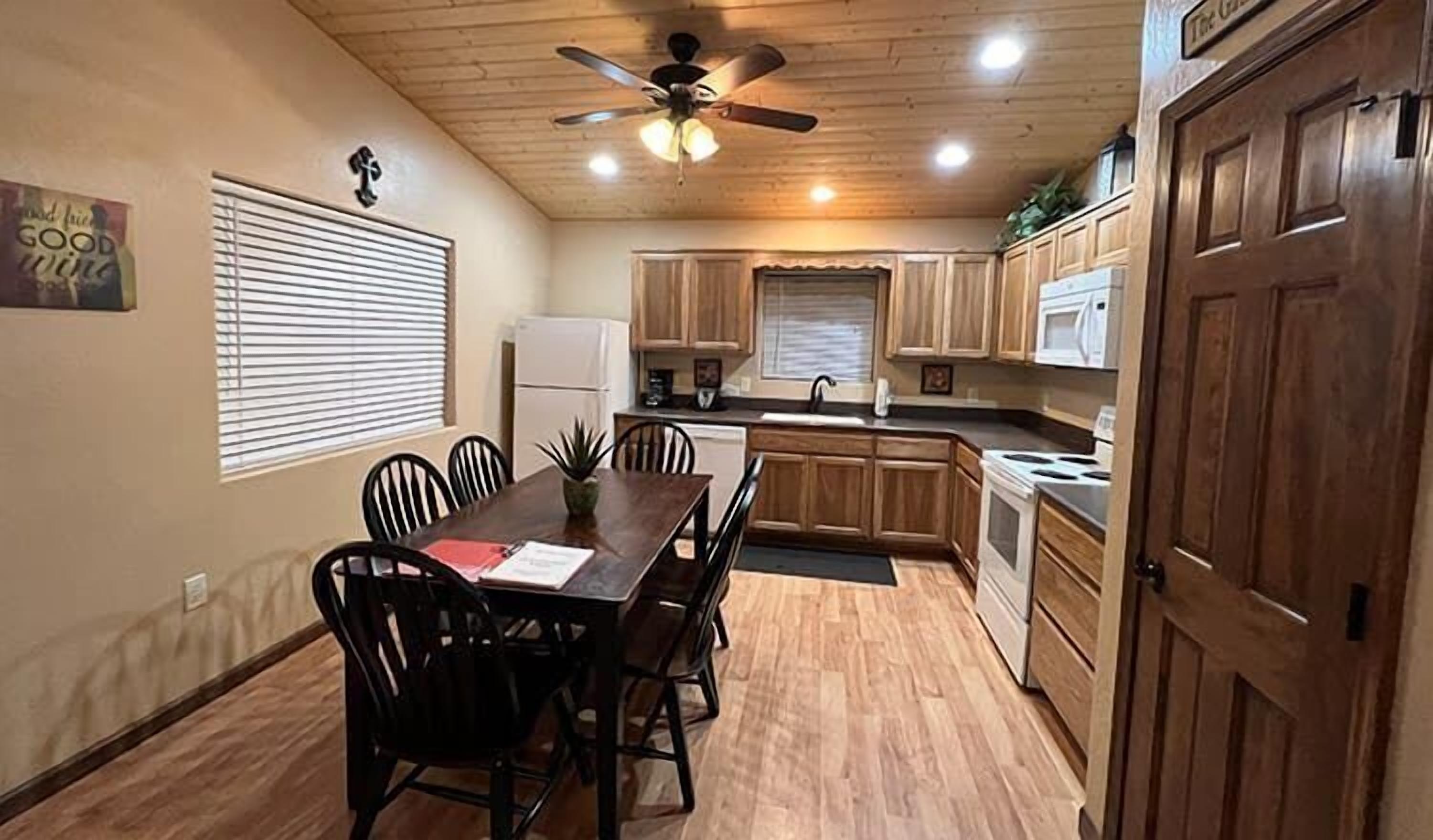 Une cuisine de taille réduite avec des meubles en bois, une table à manger avec des chaises, un ventilateur de plafond et un réfrigérateur.