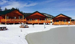 Una hilera de cabañas de madera con balcones, una mesa de picnic y un terreno cubierto de nieve.