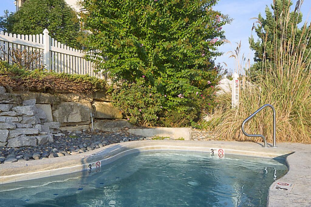 Une piscine bordée d’un muret en pierre, une clôture blanche en bois et de hautes herbes.