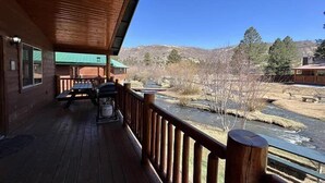 Eine Holzterrasse mit Grill und Sitzgelegenheit, mit Blick auf einen Fluss und Berge.