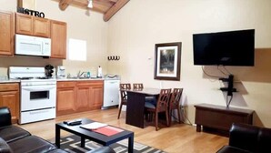 Una cocina con armarios de madera, estufa y microondas. Hay un comedor con mesa y sillas, y un televisor de pantalla plana montado en la pared.