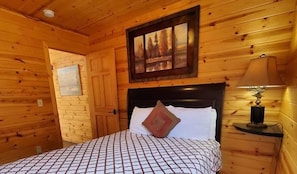 Ein Zimmer in einer Holzhütte mit einem Bett, einem Nachttisch, einer Lampe, einem Bild und einer Tür.