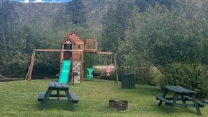 Una estructura de madera para juegos con un tobogán y columpios, rodeada de césped y árboles.