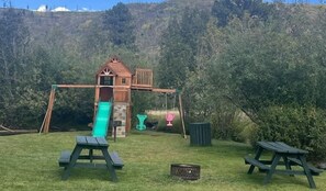 A wooden play structure with a slide and swings, surrounded by grass and trees. - Tranquil Springs Escape #94 (Greer)