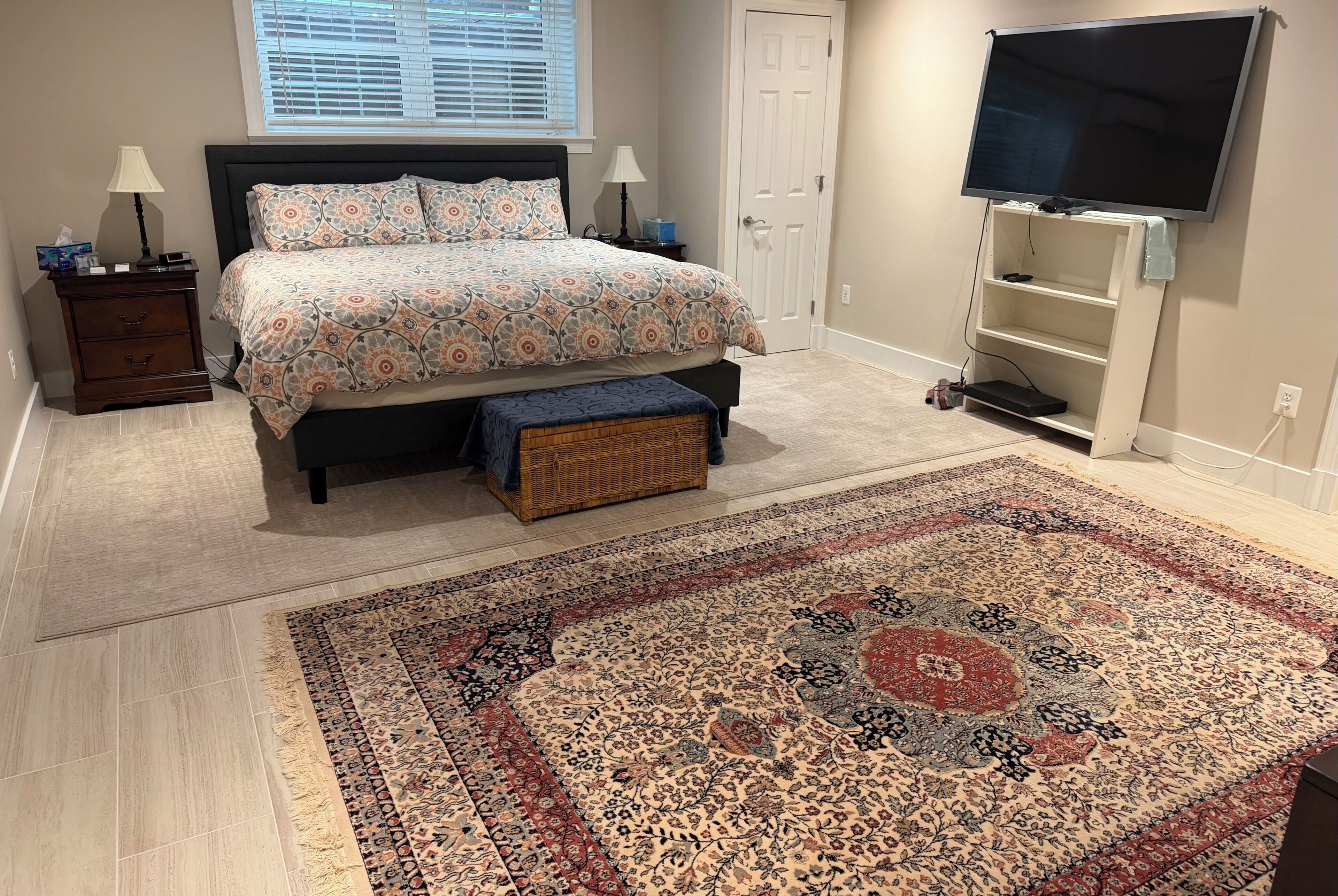 Un dormitorio con cama, mesitas de noche, televisor, puerta y alfombra estampada.